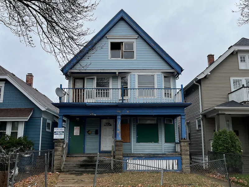 Exterior of old home with chipping blue paint, green doors, and broken windows